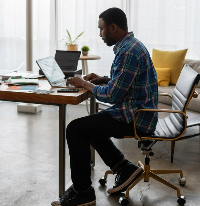 A man in a blue plaid shirt sitting at a desk typing on a laptop